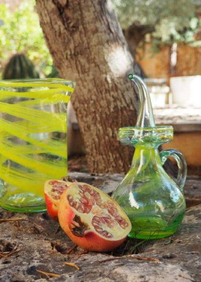 Gelber Lafiore Krug und grüne Olivenölflasche aus recyceltem Glas unter einem Olivenbaum mit aufgeschnittenem Granatapfel