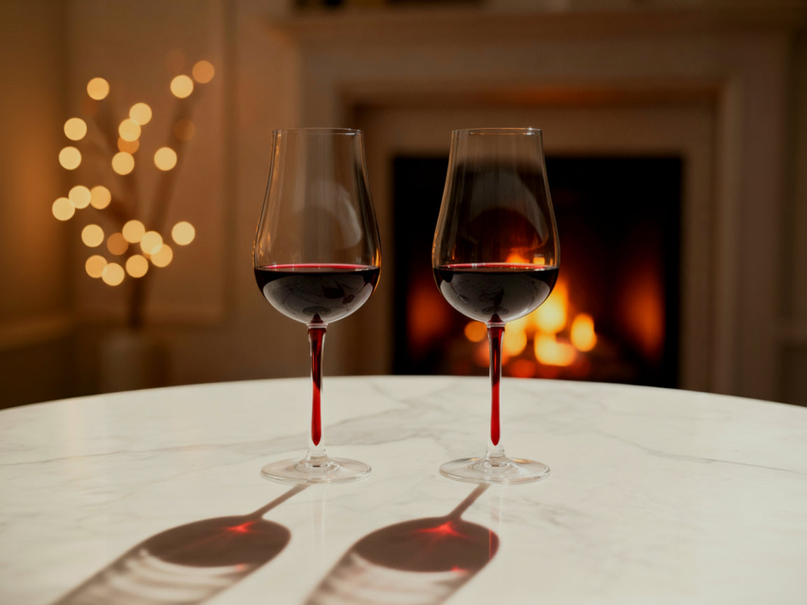Zwei gefüllte Rotweingläser mit tiefrotem Stiel auf einem Tisch vor einem Kamin